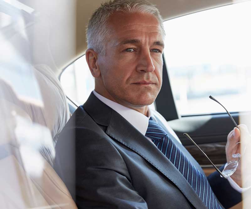 A middle-aged man in a suit and tie sits in the back seat of a car, holding eyeglasses and looking out the window with a serious expression—reflecting on strategies for Digital & Advertising Solutions.