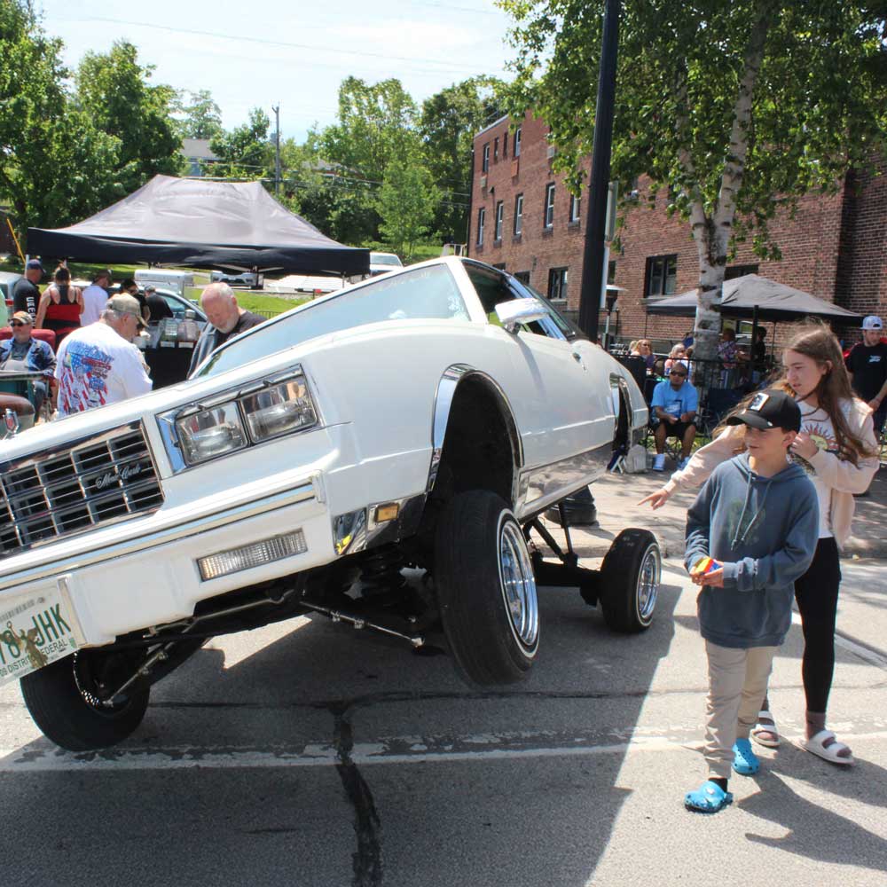 A white car with hydraulics lifts its front wheels off the ground at an outdoor event, drawing attention much like standout Print Advertising, while people watch and walk nearby.