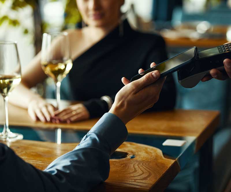 A person pays with a smartphone using contactless payment at a restaurant table, showcasing seamless Digital & Advertising Solutions, with a woman and two glasses of white wine visible.