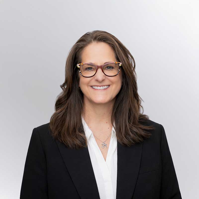 Woman with long brown hair and glasses, wearing a black blazer and white blouse, smiling in front of a plain light background—ideal for profiles on a Premier Media Network or professionals in Video & Streaming industries.