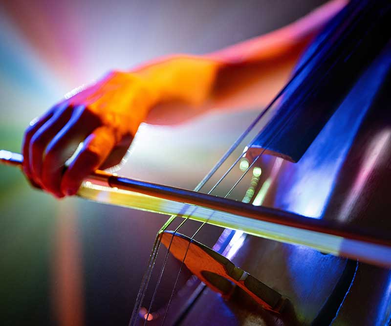 A close-up of a person's hand playing a cello with a bow, with colorful lighting in the background—perfect for showcasing Premier Media Network’s creative edge in digital & advertising solutions.