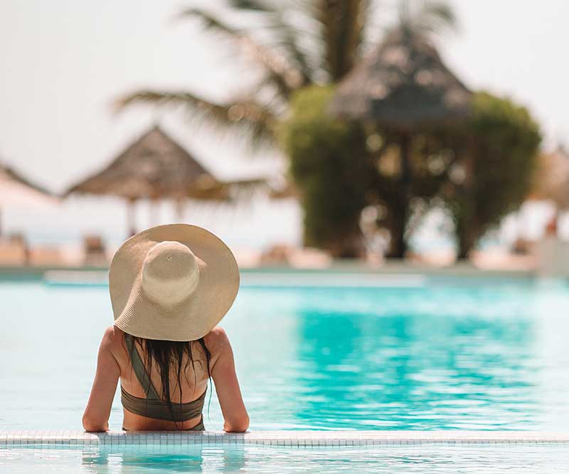 A woman in a sunhat relaxes at the edge of a swimming pool, facing away, with palm trees and beach huts in the background—a perfect scene for Digital & Advertising Solutions.