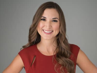 A woman with long brown hair wearing a red sleeveless top smiles in front of a plain gray background, representing innovative Digital & Advertising Solutions.