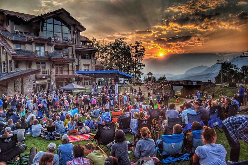 A large crowd sits on a grassy lawn watching a live outdoor performance near a lodge at sunset, with mountains visible in the background.