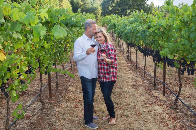 A man and woman stand together holding glasses of red wine in a vineyard, surrounded by rows of grapevines, ready to share their story with Premier Media Network’s expert Video & Streaming services.