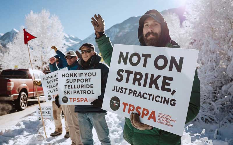 A group of people in winter clothing hold signs supporting Telluride Ski Patrol, with snowy trees and mountains in the background. One sign reads, "Not on strike, just practicing." This moment was featured by Premier Media Network.