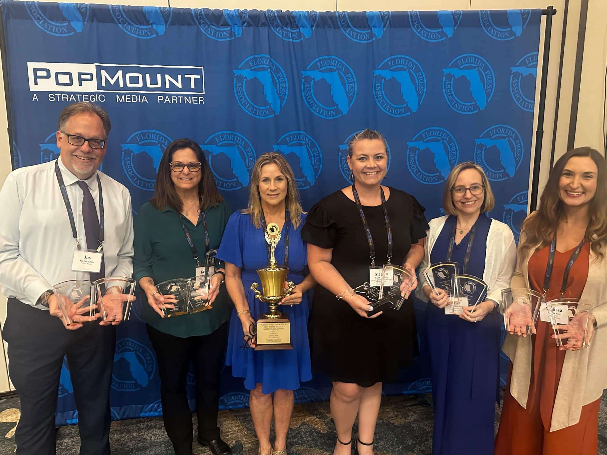 Six people stand in front of a blue backdrop holding clear glass awards and a trophy, posing for a group photo at a Premier Media Network event.