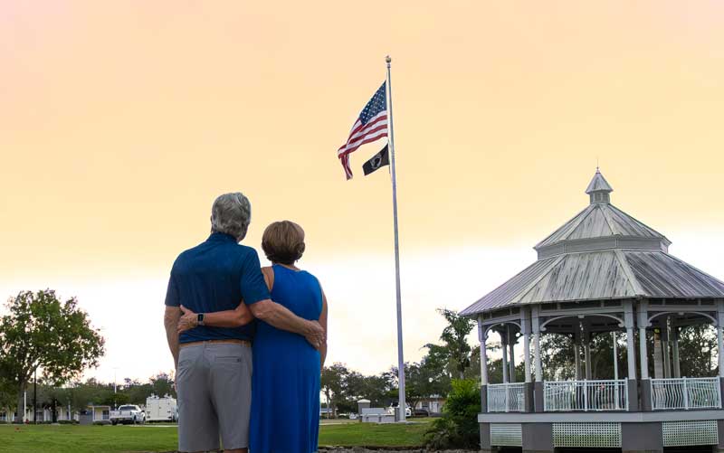An older couple stands arm in arm, facing an American flag and a gazebo at sunset—capturing a moment reminiscent of Video & Streaming campaigns, set in a serene, grassy outdoor setting.