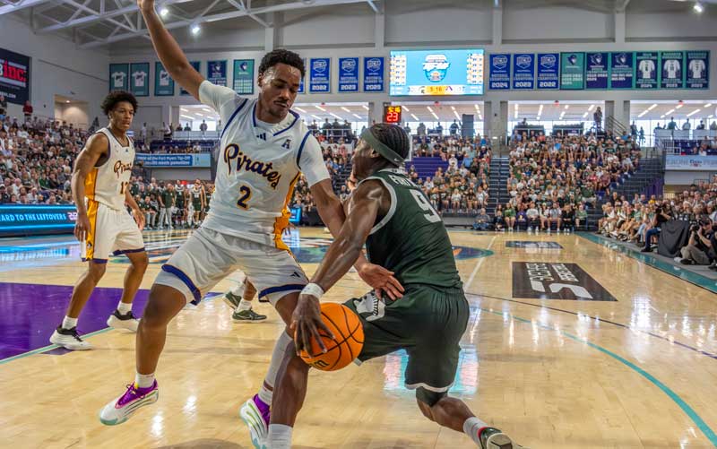 A basketball player in a green uniform drives past a defender in white during a game, as fans watch the action in a crowded indoor arena, which is being broadcast via video & streaming.
