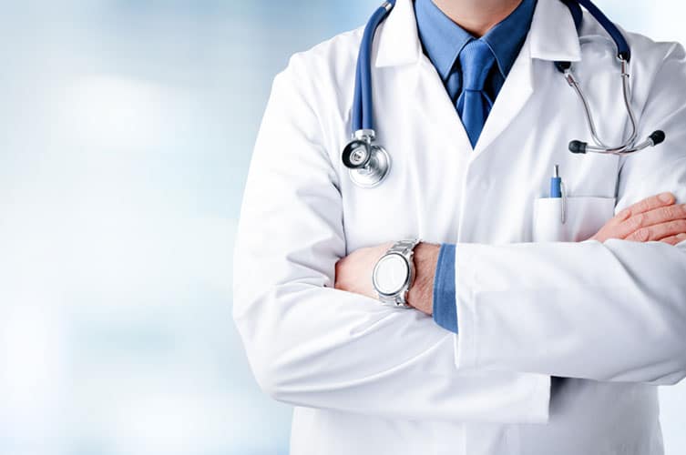 A doctor in a white coat with a stethoscope around their neck stands with arms crossed against a blurred background, embodying the trust and expertise promoted by Premier Media Network’s search engine marketing.