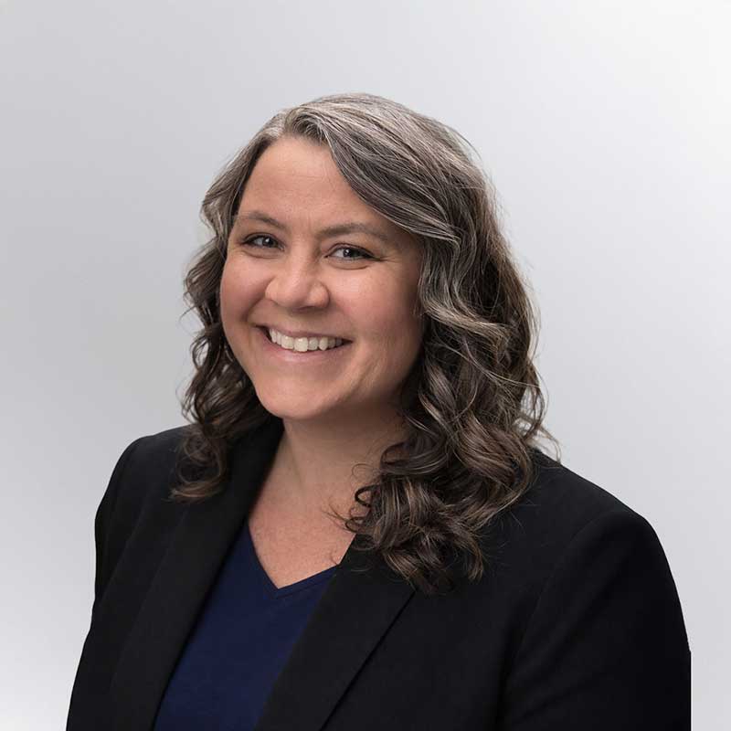 A woman with wavy brown and gray hair, wearing a black blazer and navy top, smiles at the camera against a plain light background—her expertise in programmatic advertising shines through her confident demeanor.