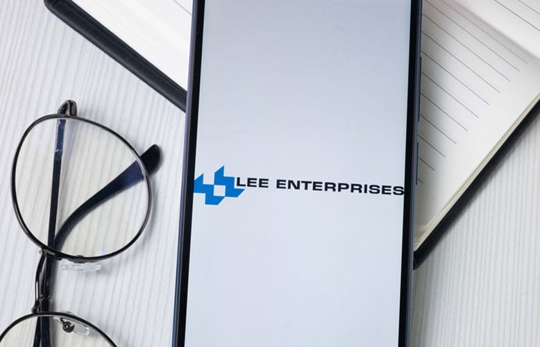 A smartphone displays the Lee Enterprises logo on its screen, highlighting a Premier Media Network, placed next to eyeglasses and an open notebook on a white surface.