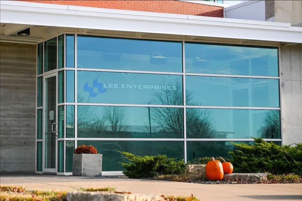 The entrance of a modern glass office building with “Lee Enterprises” on the window, three pumpkins and a planter nearby, shrubs along the walkway—home to innovative Digital & Advertising Solutions.