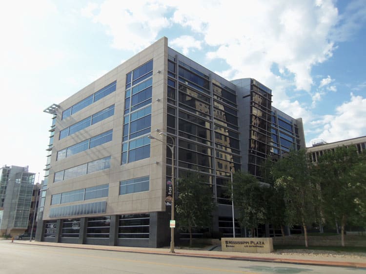 A modern multi-story office building with large glass windows displays a "Mississippi Plaza" sign, home to Premier Media Network, on a sunny day.