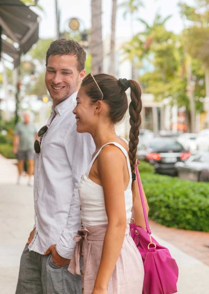 A man and woman stand on a sunny sidewalk, smiling and talking. The woman carries a bright pink bag. Both are dressed in light, casual summer clothes, perhaps discussing the latest in digital & advertising solutions from Premier Media Network.