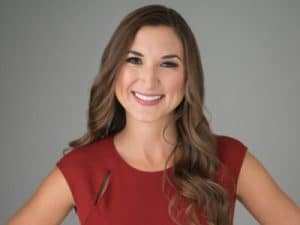 A woman with long brown hair wearing a red sleeveless top smiles in front of a plain gray background, representing innovative Digital & Advertising Solutions.