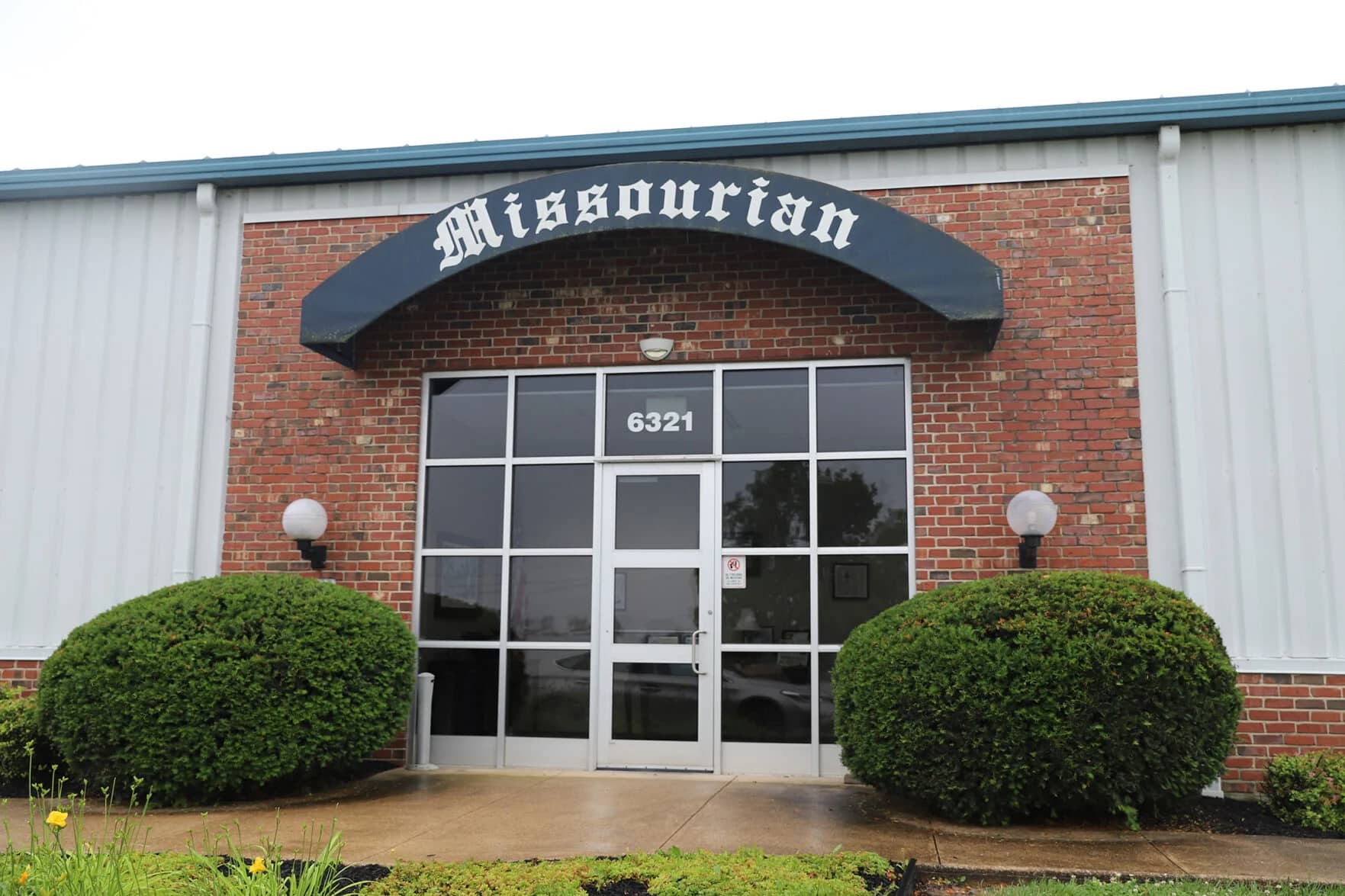 Brick building with "Missourian" sign above glass double doors, street number 6321, flanked by trimmed bushes and round outdoor lights—a hub for Digital & Advertising Solutions in the heart of the community.