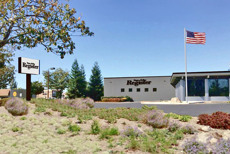 The Napa Valley Register building with sign, parking lot, and an American flag on a flagpole; landscaped greenery in the foreground—home to premier media network and digital & advertising solutions.