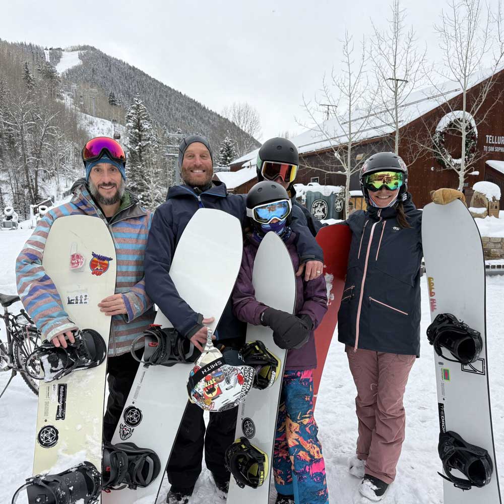 Five people in winter clothing stand together outdoors, each holding a snowboard, with snow-covered trees and a wooden building in the background—an ideal scene for showcasing digital & advertising solutions.