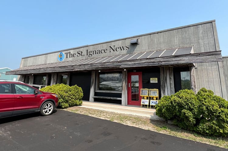 The front of The St. Ignace News building features a red door, large windows, a parked red vehicle, shrubs, and two newspaper stands by the entrance—hallmarks of a Premier Media Network dedicated to community news.
